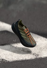 Olijfgroene sportschoen met zwarte accenten, oranje veters en drie witte strepen aan de zijkant. Zacht, flexibel materiaal en afgeronde vorm.