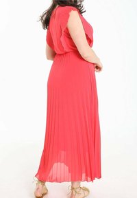 Robe longue plissée de couleur corail vif, dotée d'un col à volants et d'une silhouette fluide. Portée avec des sandales dorées à lacets.