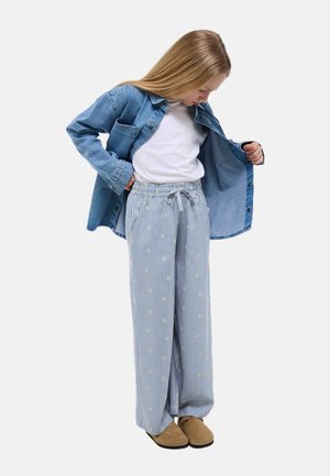 Fille portant une chemise blanche, une veste en jean bleue, un pantalon large rayé avec de petites fleurs, et des chaussures marron, regardant vers le bas en tenant sa veste ouverte.