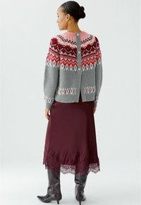 Pull en laine gris avec un motif Fair Isle coloré, col rose et fermeture à boutons au dos. Porté par-dessus une jupe en satin bordeaux avec un ourlet en dentelle.