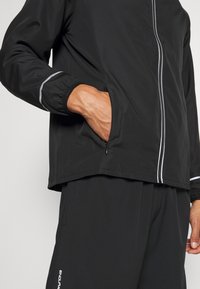 Personne portant une veste noire avec la main dans la poche zippée et un pantalon noir assorti avec un logo blanc sur la jambe gauche.
