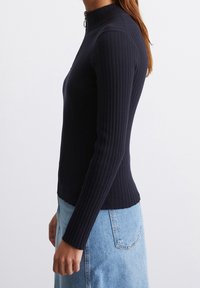 Frau in einem eng anliegenden schwarzen Rippstrickpullover mit Reißverschlusskragen und einem hellblauen Denimrock, die seitlich steht.