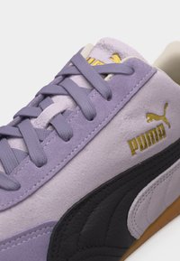 Κοντινό πλάνο ενός αθλητικού παπουτσιού Puma σε χρώμα λεβάντας με μαύρη πλευρική ρίγα, χρυσλό λογότυπο Puma και κορδόνια στο ίδιο χρώμα λεβάντας, σε λευκό φόντο.