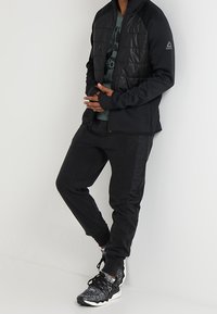 Muž v černé prošívané bundě, tmavých teplákových kalhotách a černo-bílých teniskách Reebok stojící proti bílé zdi.