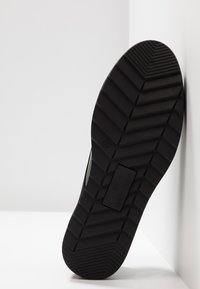 Semelle de sneaker noire avec un motif en zigzag texturé, comportant un patch logo rectangulaire. Le matériau semble être du caoutchouc avec un profil plat.