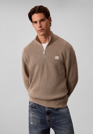 Jeune homme portant un pull beige à fermeture quart de zip par-dessus une chemise blanche et un jean bleu, debout les mains dans le dos devant un fond uni.