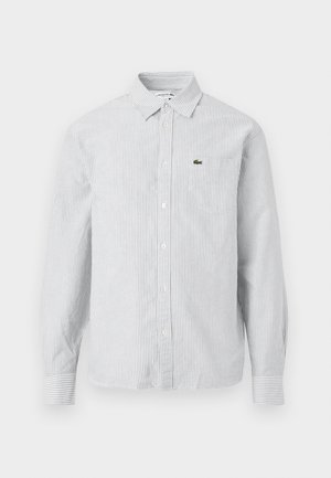 Camisa de botones a rayas en azul claro y blanco. Tela de algodón, de mangas largas, con cuello puntiagudo, que presenta un pequeño logo verde en el bolsillo del pecho.