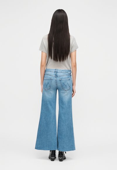 Γυναίκα με μακριά σκούρα μαλλιά, φορώντας ανοιχτό γκρι μπλουζάκι, μπλε τζιν παντελόνι με φαρδύ μπατζάκι και ραφές στις τσέπες, και μαύρα μποτάκια μέχρι τον αστράγαλο, στέκεται μπροστά σε λευκό φόντο.