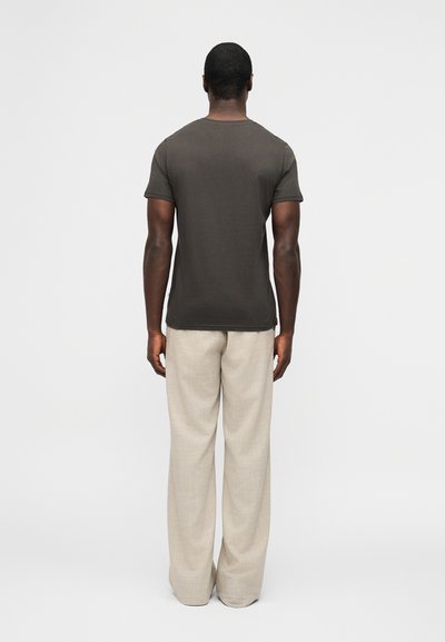 Άνδρας όρθιος με την πλάτη, φοράει εφαρμοστό μπλουζάκι σκουρόχρωμου γκρι και χαλαρό μπεζ παντελόνι από λινό, σε απλό φωτεινό φόντο.