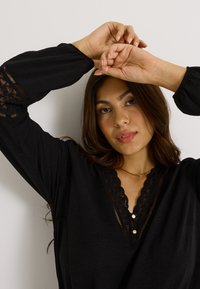 Túnica negra con mangas acentuadas en encaje y escote en V. Presenta una tela texturizada y dos botones decorativos en el cuello.