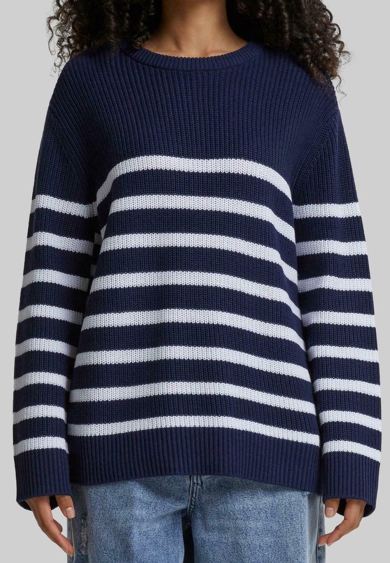 Personne portant un pull en tricot bleu marine à rayures horizontales blanches et un jean bleu, se tenant devant un arrière-plan uni.