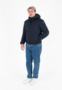 Granatowa kurtka puchowa z kapturem, z przednim zamkiem, kieszenią na piersi i kontrastującymi żółtymi akcentami, zestawiona z niebieskimi dżinsami i białymi trampkami.