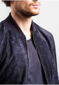 Námornícka zamšová bomber bunda s rebrovaným golierom, predným zipsom a textúrovaným povrchom; preložená cez tmavú košeľu. Detaily látky a blízky pohľad.