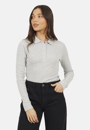 Jonge vrouw met lang bruin haar, gekleed in een lichtgrijs poloshirt met lange mouwen en zwarte broek met hoge taille, met één hand in de zak.