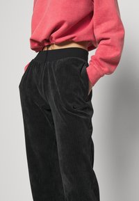 Black corduroy pants with a relaxed fit, featuring vertical stripes and side pockets. The waistband is elastic, paired with a pink sweatshirt.