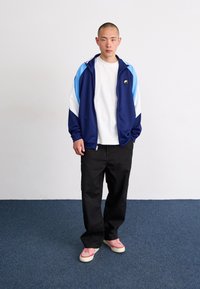 Casaco desportivo em azul marinho e azul claro com detalhes brancos, combinado com uma t-shirt branca e calças pretas. Ténis rosa completam o look.