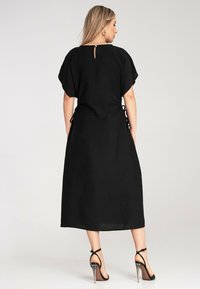 Schwarzes Maxikleid mit lockerer Passform, kurzen Ärmeln und einem Ausschnitt im Rücken. Mit Schnürdetails an den Seiten und einer glatten Textur.