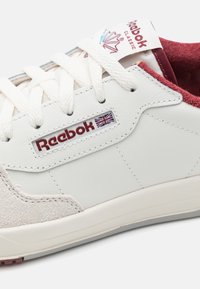 Valkoiset Reebok-kengät, joissa on nubukkiyksityiskohtia, tekstiili- ja nahkapintaa, näkyvä logo sekä maroonit yksityiskohdat kielekkeessä ja kaulassa. Klassinen nauhakäyttöinen muotoilu.