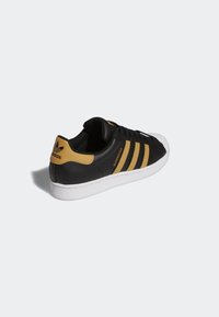 Black leather sneaker with three horizontal gold stripes, white rubber sole, and textured black accents. Branding on the heel.