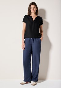 Haut noir à manches courtes avec un col en V, accompagné d'un pantalon large bleu marine et de baskets blanches avec des accents rouges, sur un fond neutre.