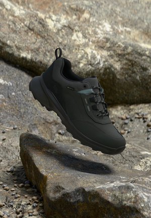 Chaussure de randonnée noire avec étiquette Gore-Tex suspendue au-dessus d'un terrain rocheux, montrant la semelle et les lacets sur un fond de pierre texturée.