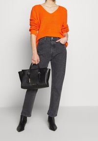 Orange ribbad v-ringad tröja, grå högmidjade jeans, svart läderhandväska och svarta ankelskor på en neutral bakgrund.