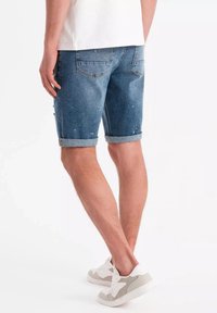 Jeansshorts i blått med en blekt tvätt, uppvikta ärmslut, två bakfickor och subtila färgstänk. Kombinerade med vita och grå sneakers.