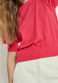 Růžový pletený pulovr s krátkými rukávy a žebrovaným lemem. Měkká textura, volný střih, nošený s světlými kalhotami.