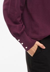 Paarse gebreide trui met een aansluitend ontwerp en gestructureerde mouwen, gedetailleerd met drie parelknopen op de manchetten, gecombineerd met zwarte broek.