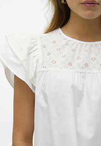 Donna che indossa una blusa bianca con maniche a sbuffo e un dettaglio in pizzo floreale sullo scollo, vista dalle spalle fino a metà torace.