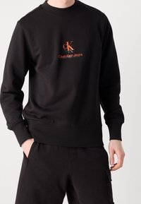 Černá mikina s kulatým výstřihem, žebrovanými manžetami a lemem. Na hrudi je vyšívané logo Calvin Klein Jeans v červené a oranžové barvě.