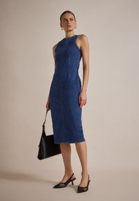 Rochie din denim albastru, ajustată pe corp, cu design fără mâneci, detaliu cu cusătura frontală, lungime până la genunchi, combinată cu pantofi slingback negri și o poșetă.