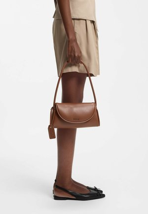 Person wearing beige shorts and black flats holds a brown leather Hugo handbag with a shoulder strap and branded tag.