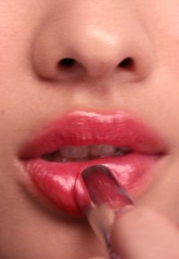 Rossetto applicato su labbra piene, lucide e rosa; lo strumento per l'applicazione è mostrato mentre tocca la superficie delle labbra; una leggera lucentezza è visibile.