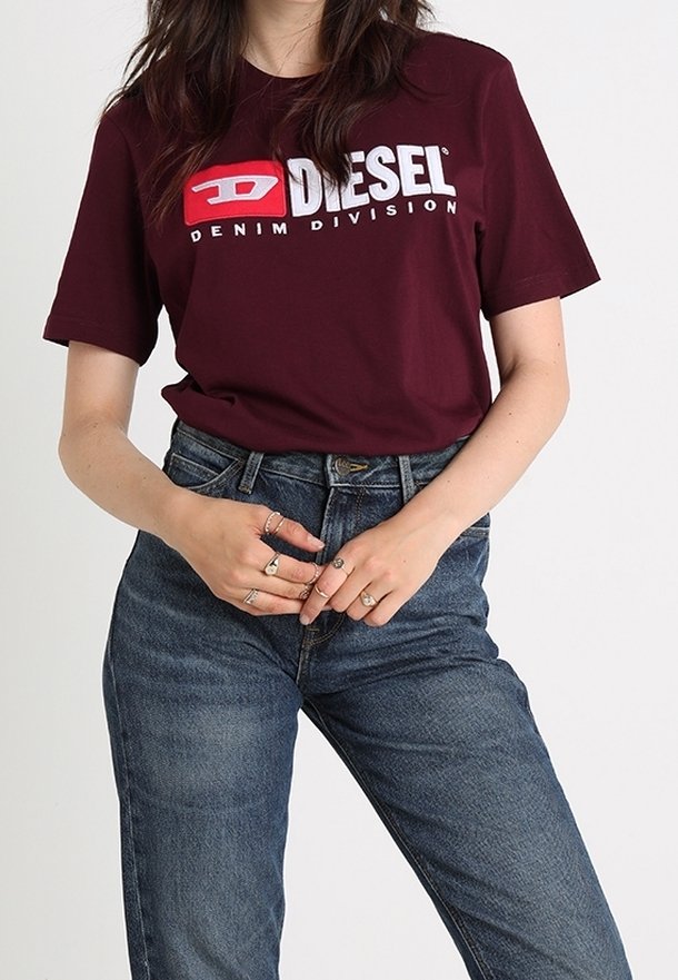 Burgunderfärgad kortärmad t-shirt med framträdande vit "DIESEL"-logotyp och texten "DENIM DIVISION", i kombination med blå denimjeans.