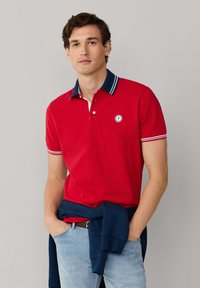 Hombre de pie con las manos en los bolsillos, vistiendo una camisa polo roja con cuello azul marino y pantalones vaqueros azul claro, con una sudadera azul marino atada alrededor de la cintura.