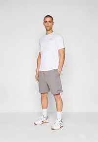 Valge lühikeste varrukatega sportlik särk, hallid lühikesed püksid külgedel taskutega, valged tossud mustade aktsentidega, seismas ühtlase tausta ees.