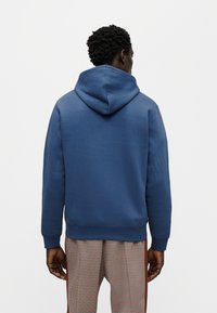 Μπλε φούτερ με κουκούλα κατασκευασμένο από μαλακό ύφασμα, με τσέπη καγκουρό και ριμπ μανσέτες, εμφανιζόμενο από πίσω, με καφέ και πορτοκαλί καρό παντελόνια.