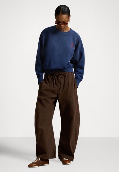 Marineblå genser med en rød logo, kombinert med løstsittende brune bukser og brune snøresko. Avslappet og komfortabel passform.