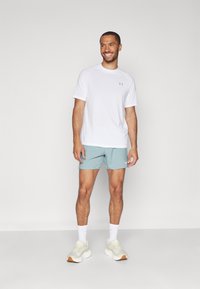 Camiseta atlética blanca de manga corta con un logo gris, combinada con pantalones cortos azul claro. Lleva calcetines blancos y zapatillas deportivas crema con suelas marrones.