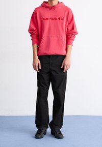 Felpa con cappuccio rossa con logo ricamato, tasca frontale e lacci, abbinata a pantaloni neri e sneaker nere su un pavimento blu.