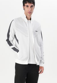 Bela zip-up jakna z sivo podrobnostjo, teksturiran vzorec spredaj in stoječim ovratnikom, v kombinaciji z belo majico pod njo.