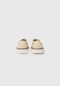 Beige sneakers with a textured upper, contrasting brown sole, and metal accents. Features a rounded shape and visible stitching detail.