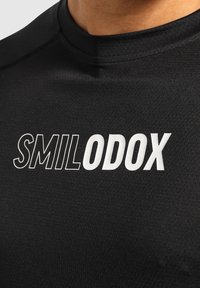 Černé sportovní tričko s texturovanou tkaninou a kulatým výstřihem. Na přední straně je zobrazeno jméno značky "SMILODX" tučným bílým písmem.
