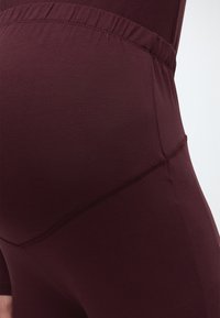 Lähivõte inimesest, kes kannab tumeburgundia värvi rasedate leggings'eid kõrge elastse vöökoha ja nähtavate õmblustepoltega.