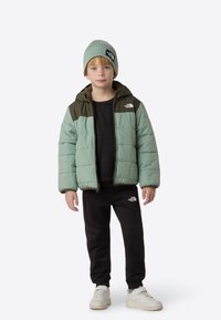 Heleroheline ja tumeoliiviroheline kampsunjope koos kapuutsi ja eesmise tõmblukuga. Sobitatud musta dressipluusi ja dressipükstega ning valgete spordijalatsitega.