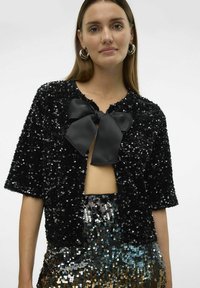 Chaqueta corta de lentejuelas negras con mangas cortas y cuello redondo, que presenta un gran lazo de satén negro en la parte delantera. Textura brillante.