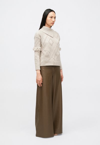 Nainen seisoo päällään beige, teksturoitu neule, jossa on hapsureunus, ja leveälahkeiset ruskeat housut, valkoista taustaa vasten.