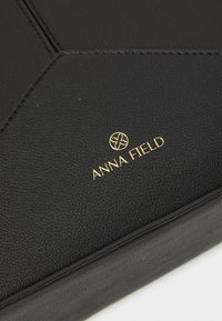 Surface en cuir texturé noir avec des détails cousus et le nom de la marque "Anna Field" ainsi que le logo dorés centrés sur le matériau.