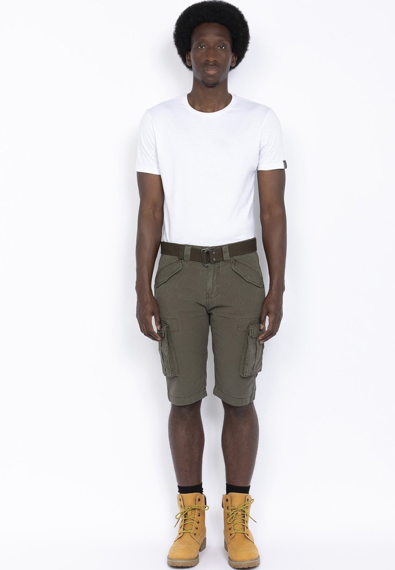 Pantaloni cargo verde oliva con tasche laterali, abbinati a una t-shirt bianca aderente e stivali gialli. Una cintura marrone mette in risalto la vita.